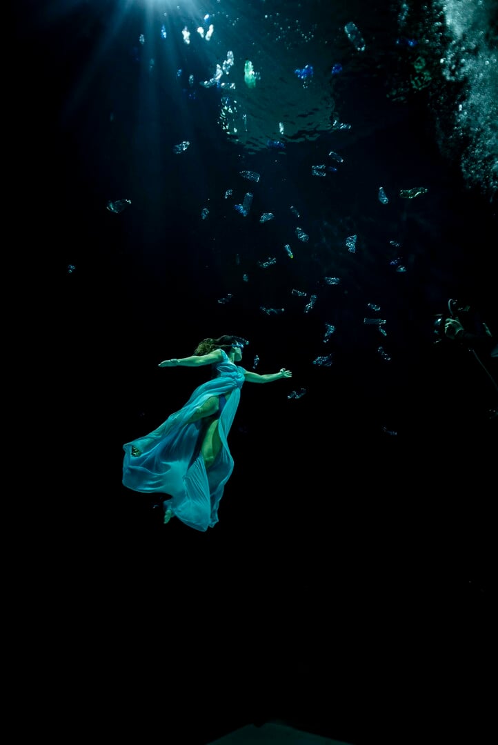A woman wearing a flowing dress floats in a black abyss with her hand stretched out towards a cascade of trash sinking towards the bottom.