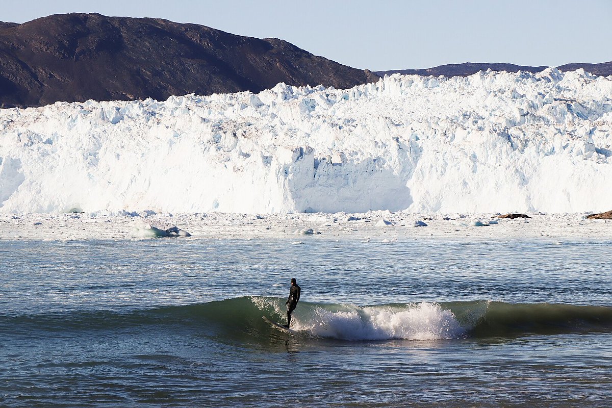 A surfer rides a wave with melting glaciers in the background
