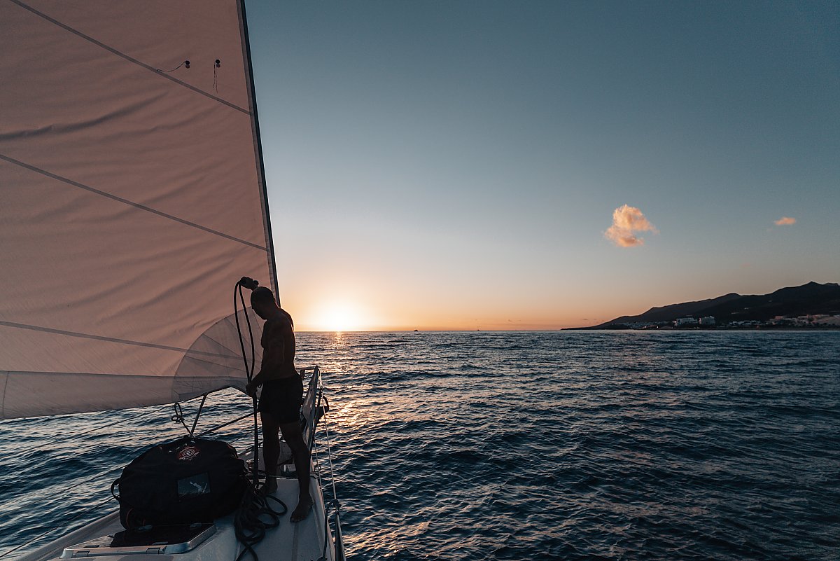 Man works on rigging on a sail boat out in on the ocean as the sun sets/rises.