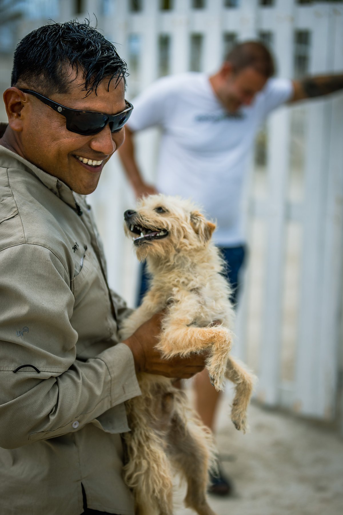 Smiling man wearing dark sunglasses holds a scruffy medium size dog with beige fur. Another man out of focus stands in the background.