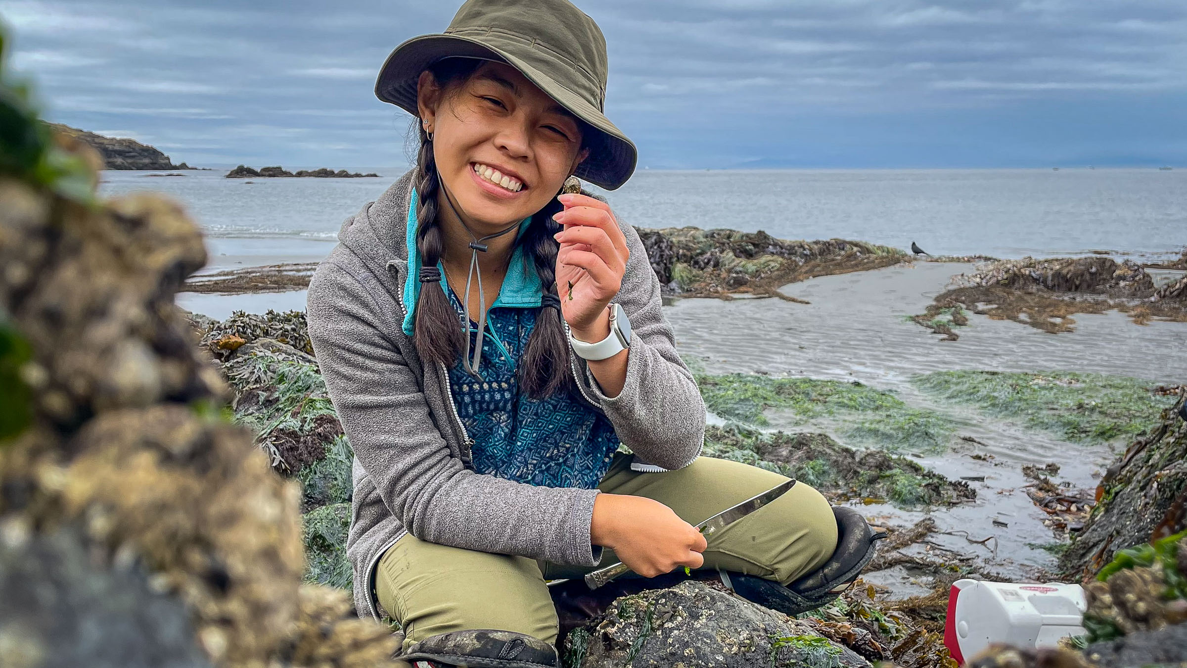 a person in a hat smiling while sitting on a rock near shore